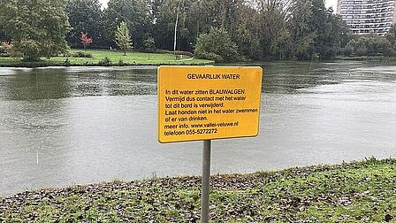 Waarschuwingsbord met de tekst 'GEVAARLIJK WATER. In dit water zitten BLAUWALGEN. Vermijd dus contact met het water tot dit bord is verwijderd. Laat honden niet in dit water zwemmen of ervan drinken. Meer info: Vallei-veluwe.nl. Telefoon: 055 - 5272272